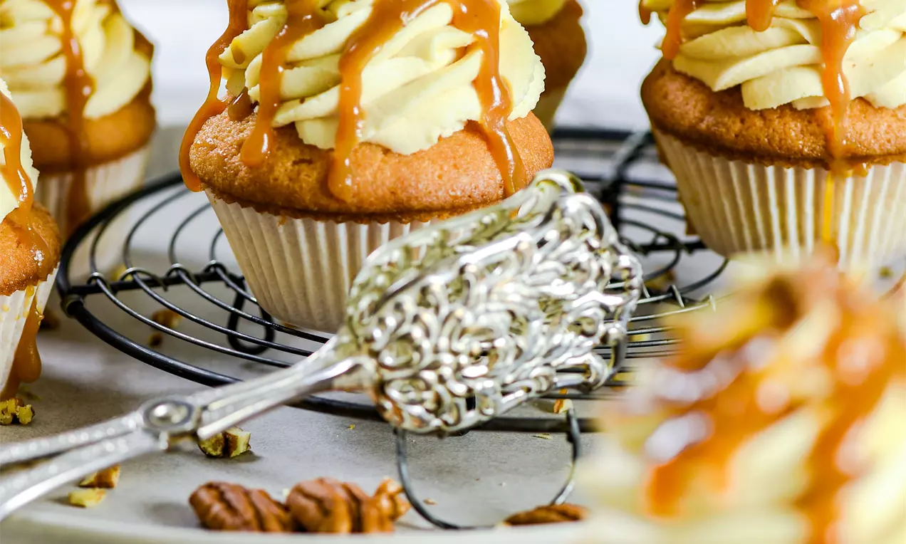 Picture - Lysebrune cupcakes med kremhvit topping, dryppet med gyllen karamell og dryss av grovhakkede pekannøtter på et metallgitter