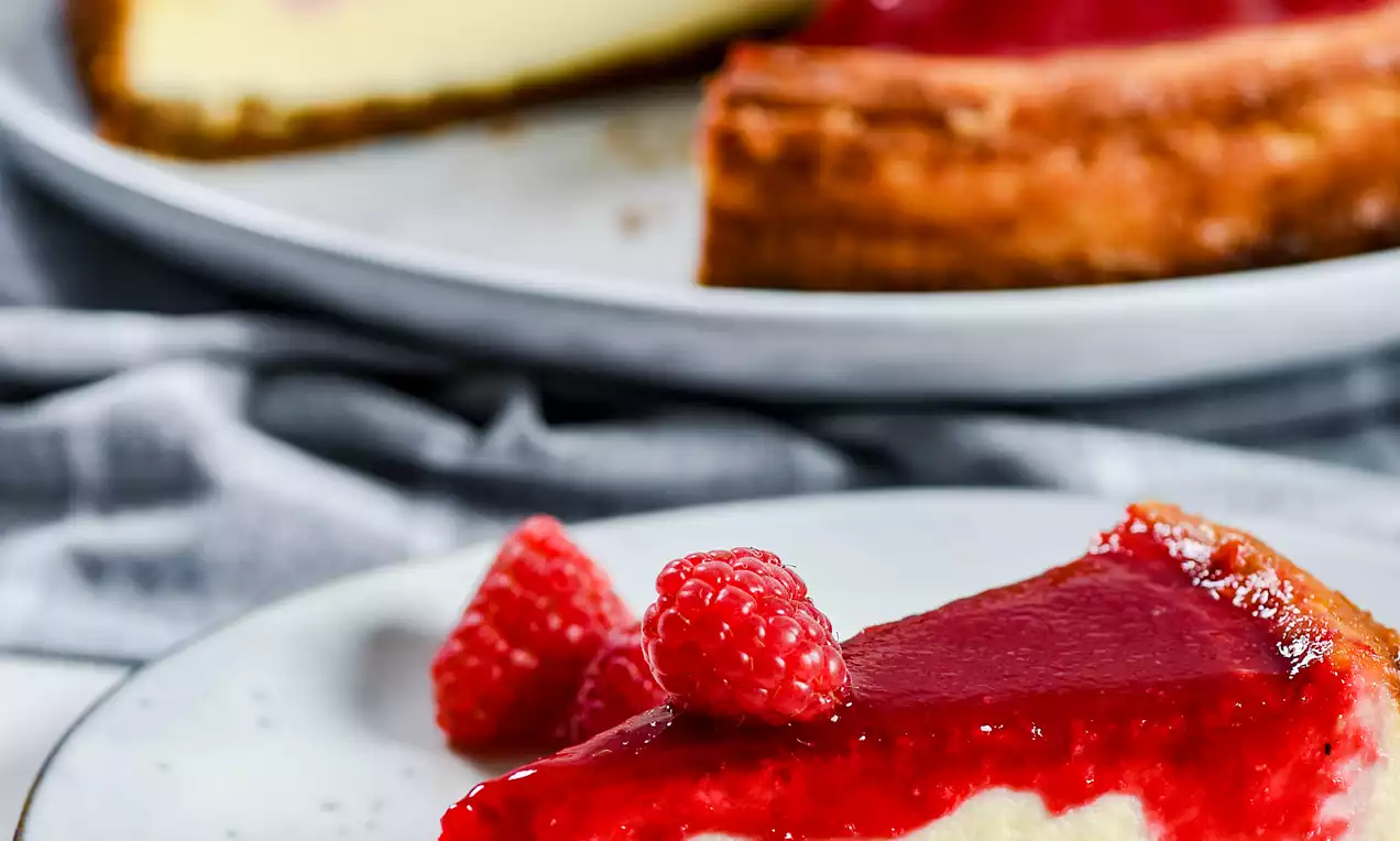 Picture - Baked raspberry cheesecake med gyllenbrun kjeksbunn, hvitt kremfyll og blank, rød bringebærgele toppet med friske bringebær på hvit tallerken