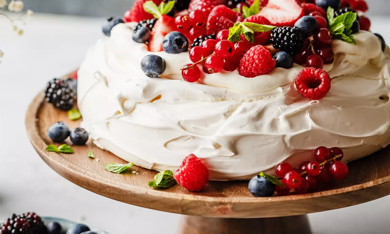 Picture - Vaniljefarget Pavlova med myke marengsbølger, toppet med røde og lilla bær, mynteblader og plassert på et rustikt trefat