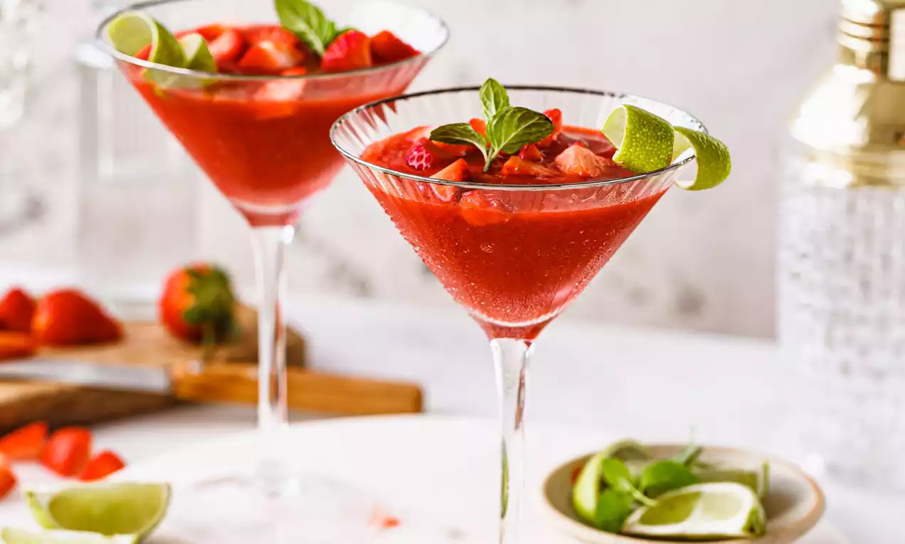 Picture - To høye cocktailglass med rød Strawberry Vanilla Daiquiri, pyntet med limebåter, jordbærbiter og mynteblad, på lys marmorfat